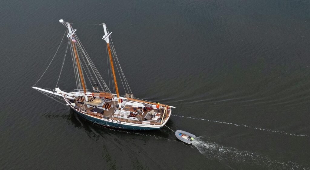 Aerial view of a classic wooden sailboat towing a small motorboat across calm water