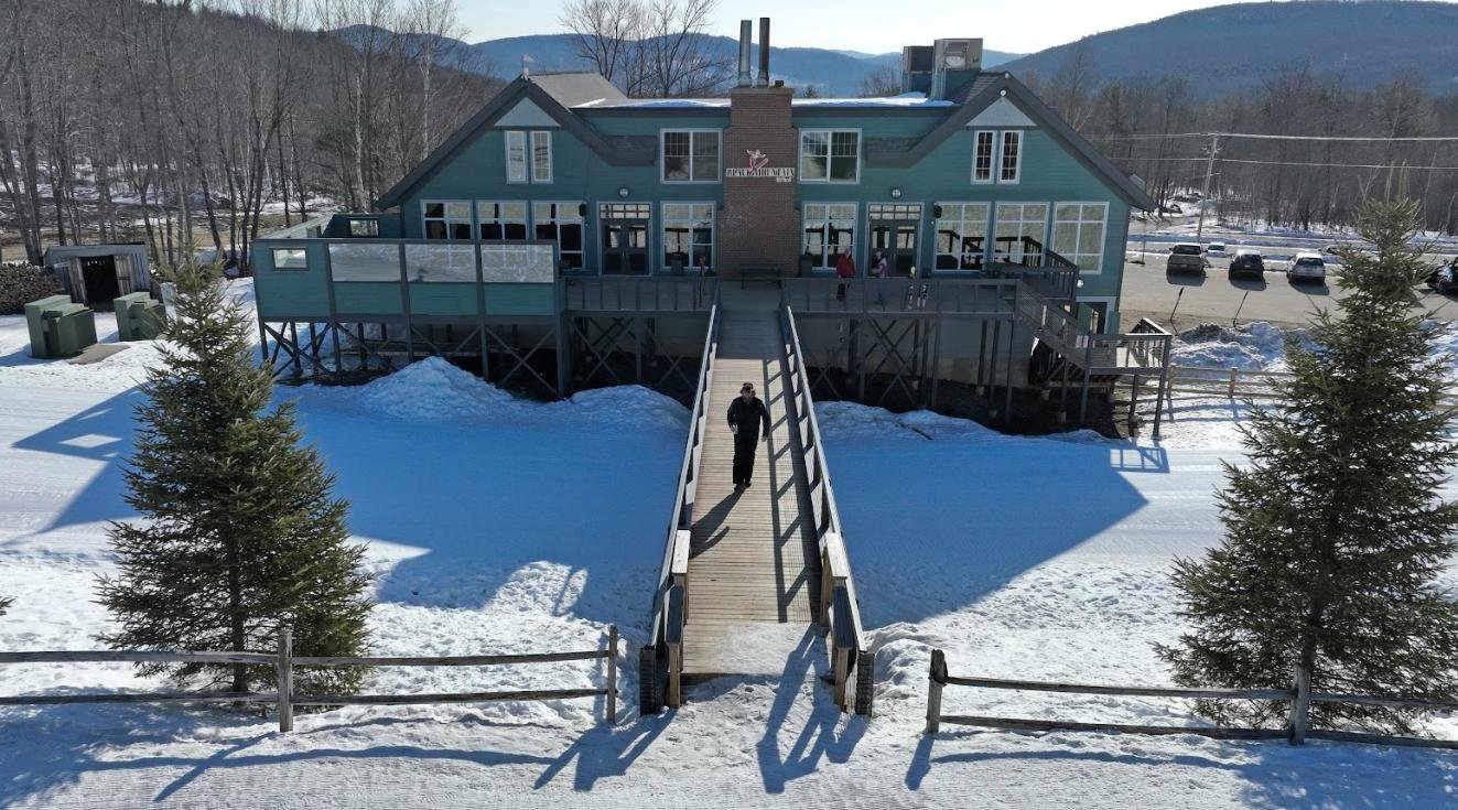 Aerial view of a person walking up a wooden ramp toward a mountain lodge surrounded by snow and evergreen trees.