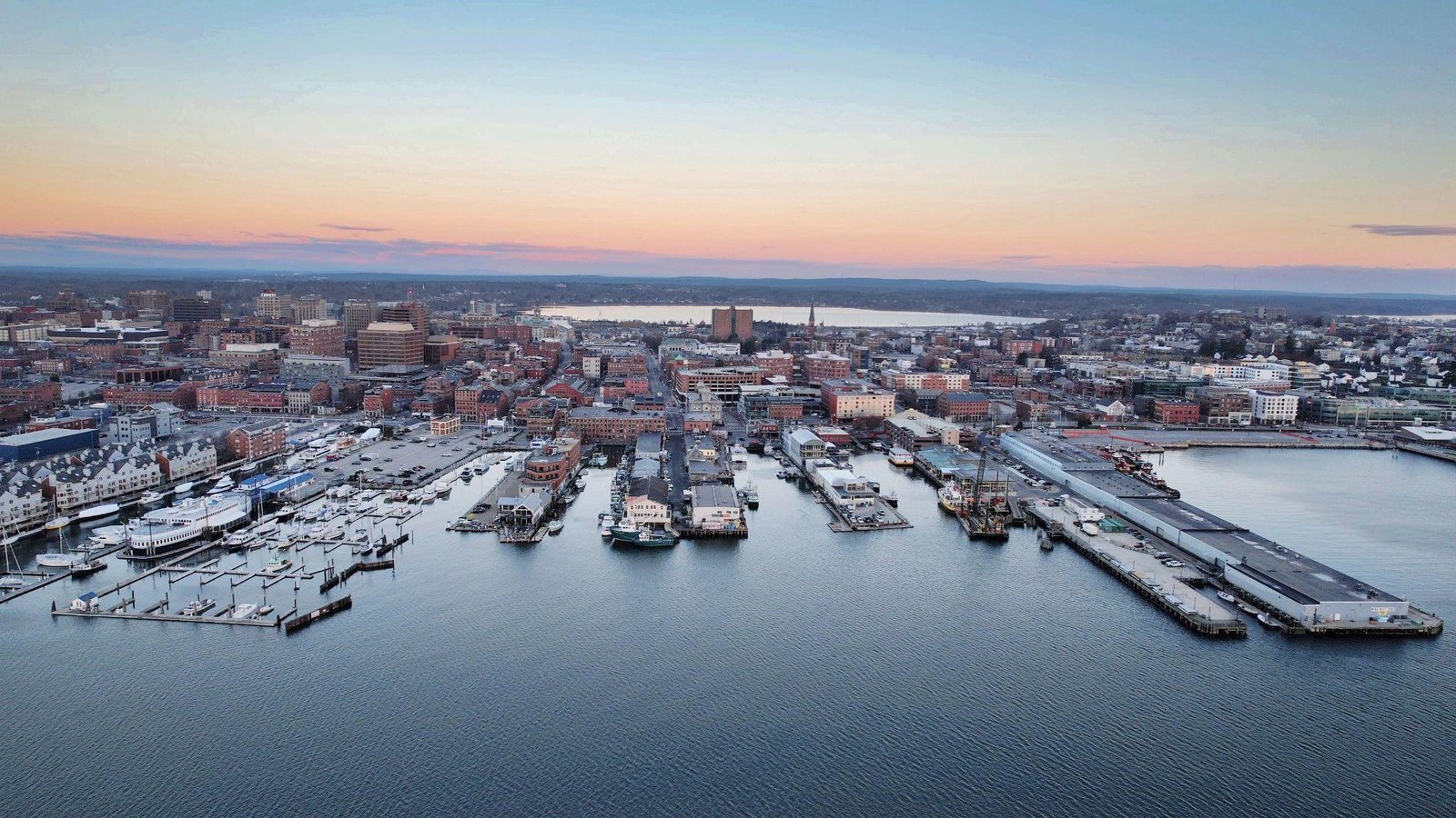 Aerial drone view of Portland, Maine waterfront at sunset with marina, fishing piers, and city skyline.