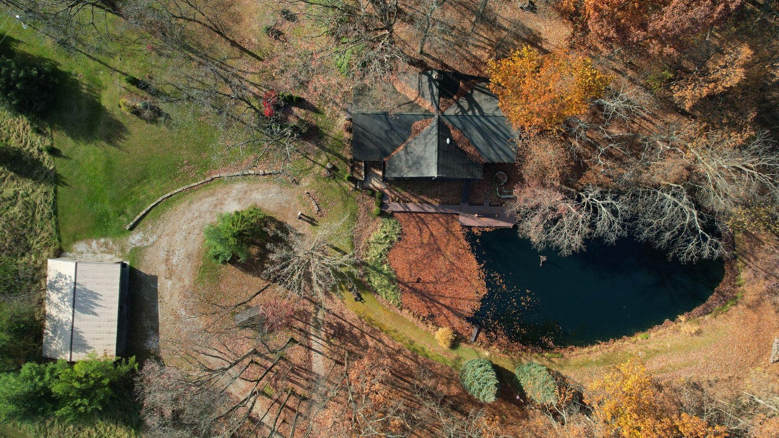 Aerial drone view of a rural property with a cabin, private pond, outbuilding, and autumn woodland surroundings.